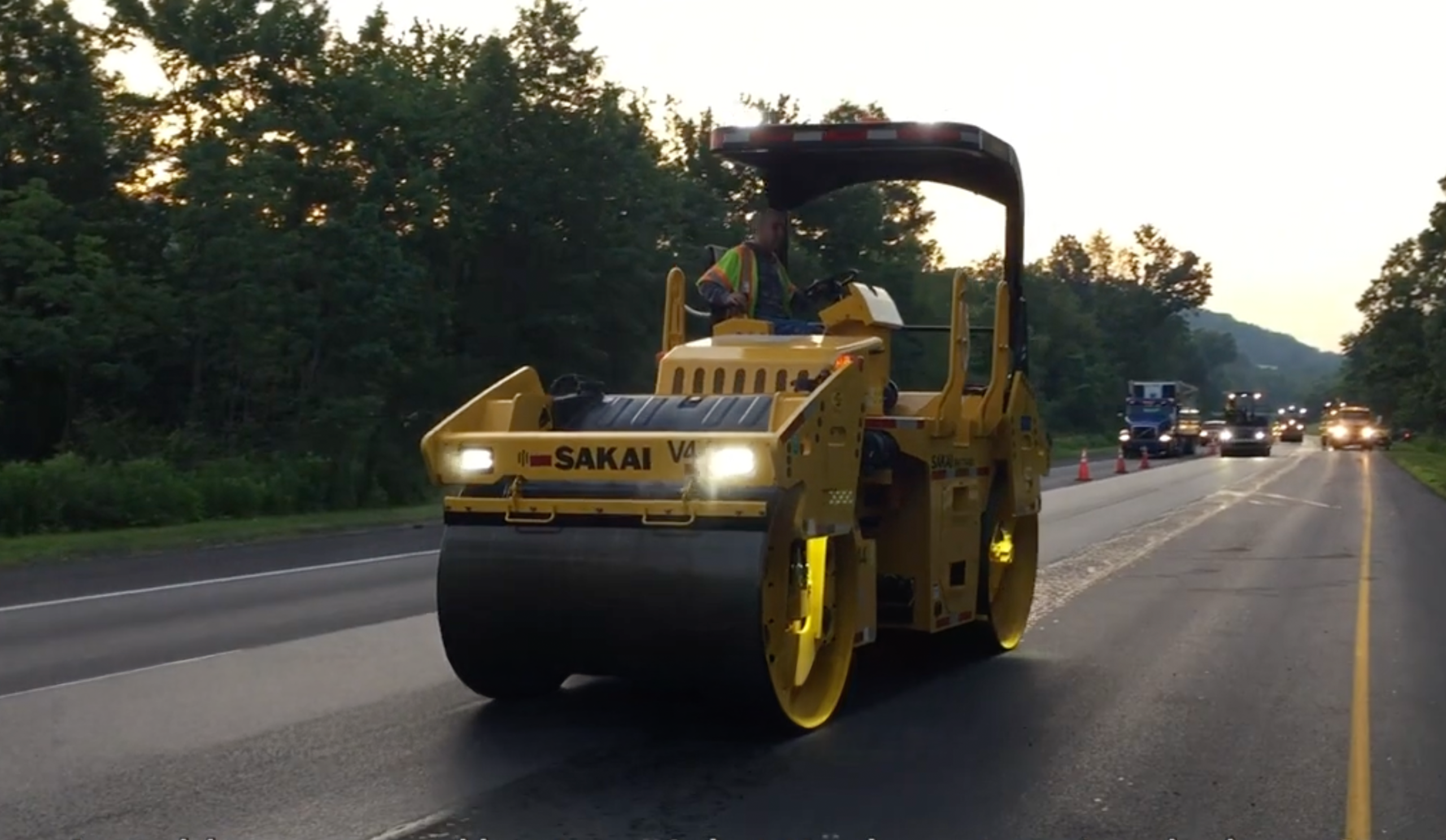 Sakai roller with headlights on, paving a road at dusk