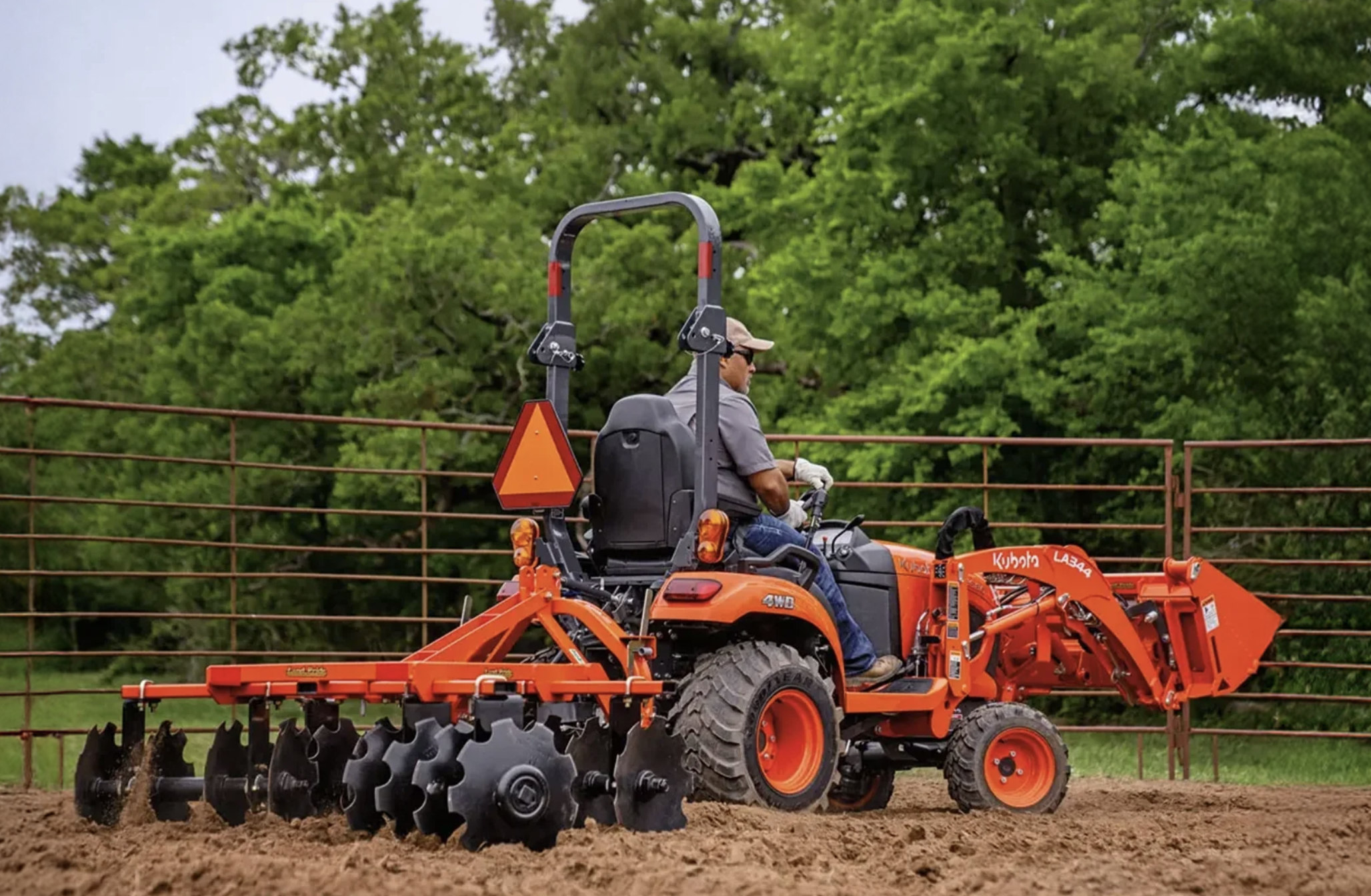 Operator driving a Kubota tractor with a front loader and a rear implement across grass